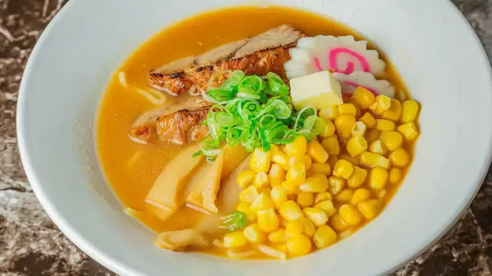 Tonkotsu Ramen is a bowl of ramen in a rich, creamy pork bone broth, topped with grilled pork slices, corn, green onions, bamboo shoots, and a decorative fish - cake slice at Kokoro Sushi, a Japanese restaurant in CA